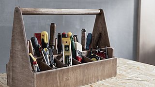 Foto, ein hözerner Werkzeugkasten auf einem Tisch. In dem Werkzeugkasten befinden sich viele verschiedene Werkzeuge.